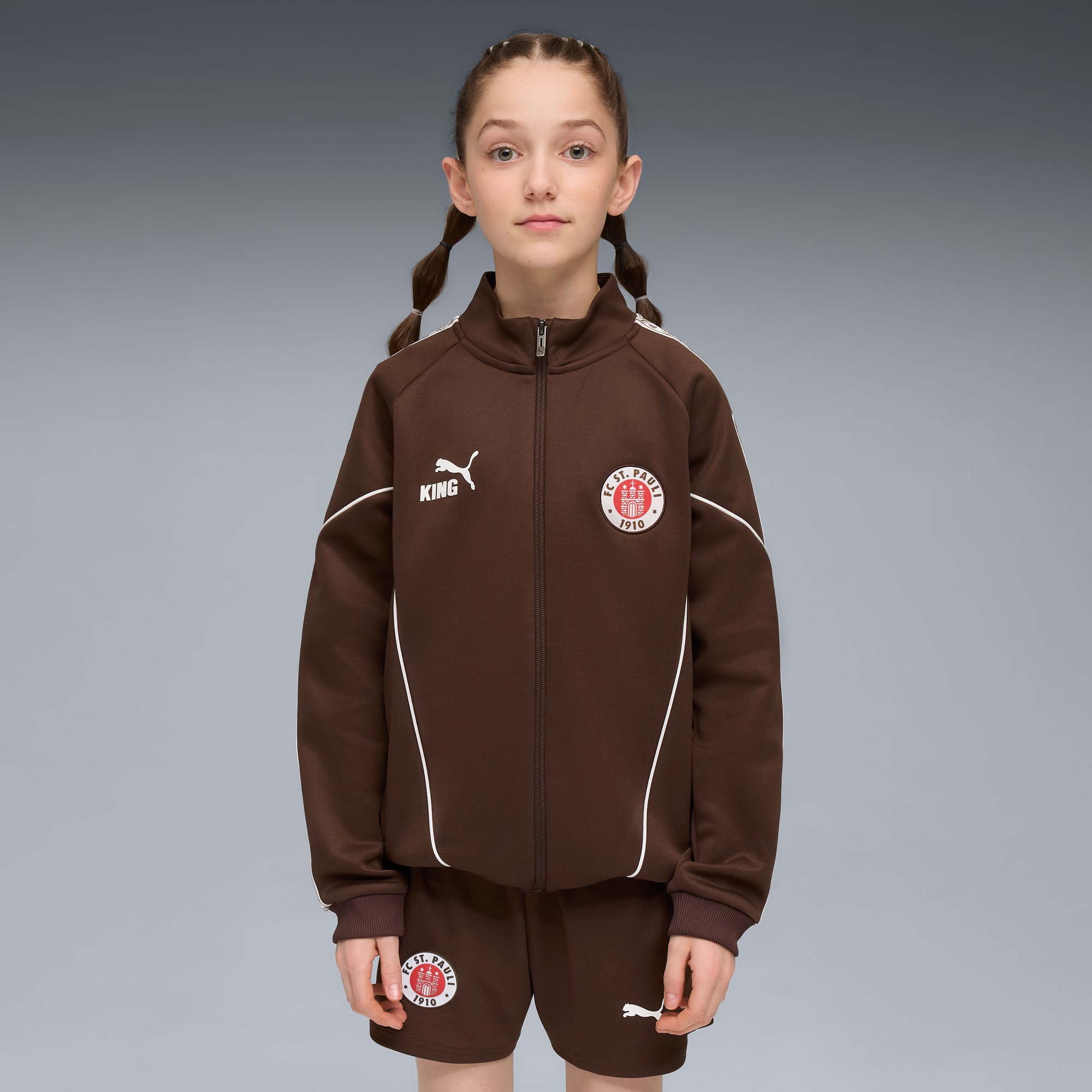 Sur fond gris, un mannequin pose en veste marron et short avec emblèmes d'équipe.